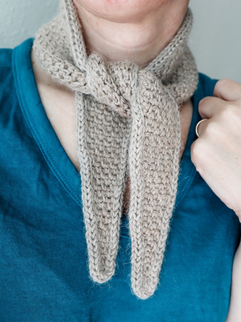 K&ouml;nnte beinhalten: Ein beigefarbener H&auml;kel-Schal, der um den Hals einer Person gebunden ist. Der Schal ist aus weichem, flauschigem Garn gefertigt und hat ein schlichtes, elegantes Design.