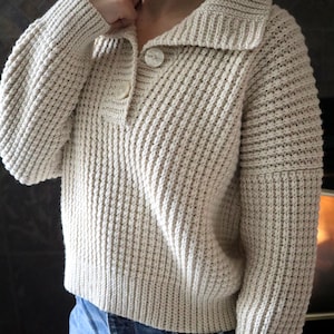 May include: A cream-colored knitted sweater with a collared neckline and two button closures. The sweater has a waffle knit texture and long sleeves.