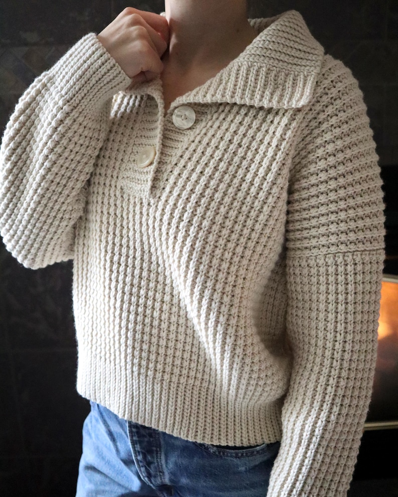 May include: A cream-colored knitted sweater with a collared neckline and two button closures. The sweater has a waffle knit texture and long sleeves.