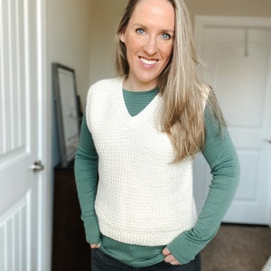 May include: A woman wearing a white knitted vest over a teal long-sleeved shirt and black jeans.