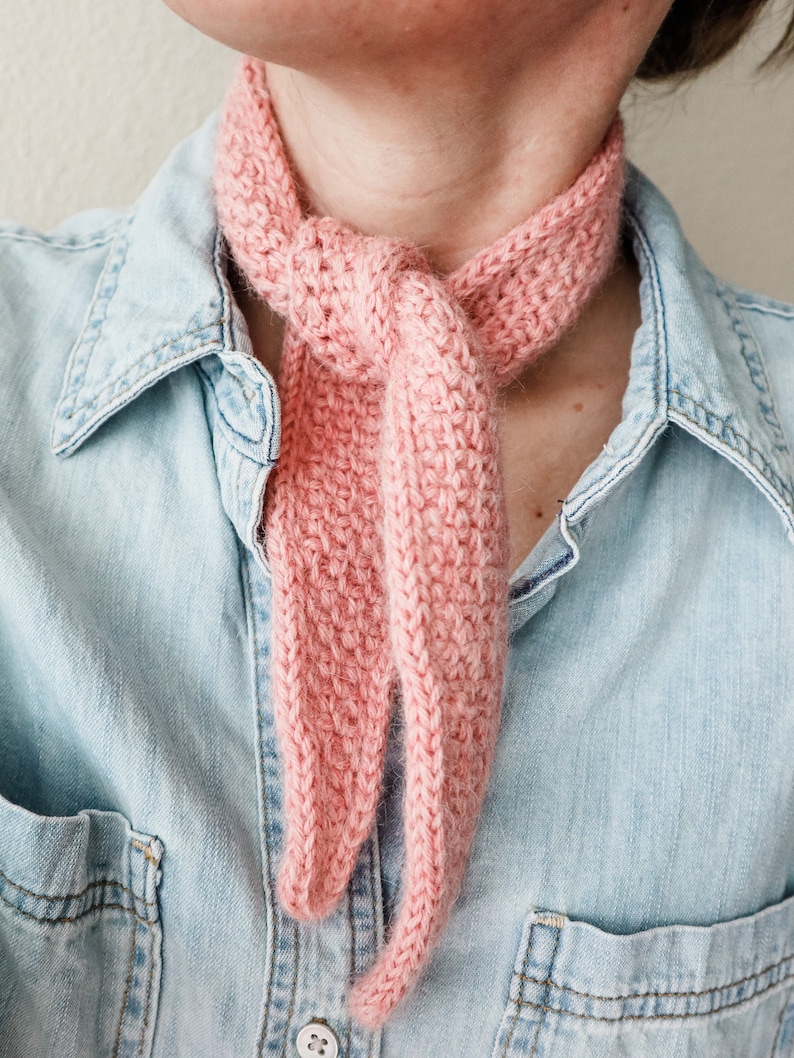 K&ouml;nnte beinhalten: Ein rosa geh&auml;kelter Schal, der um den Hals einer Person gebunden ist, die ein hellblaues Jeanshemd tr&auml;gt.