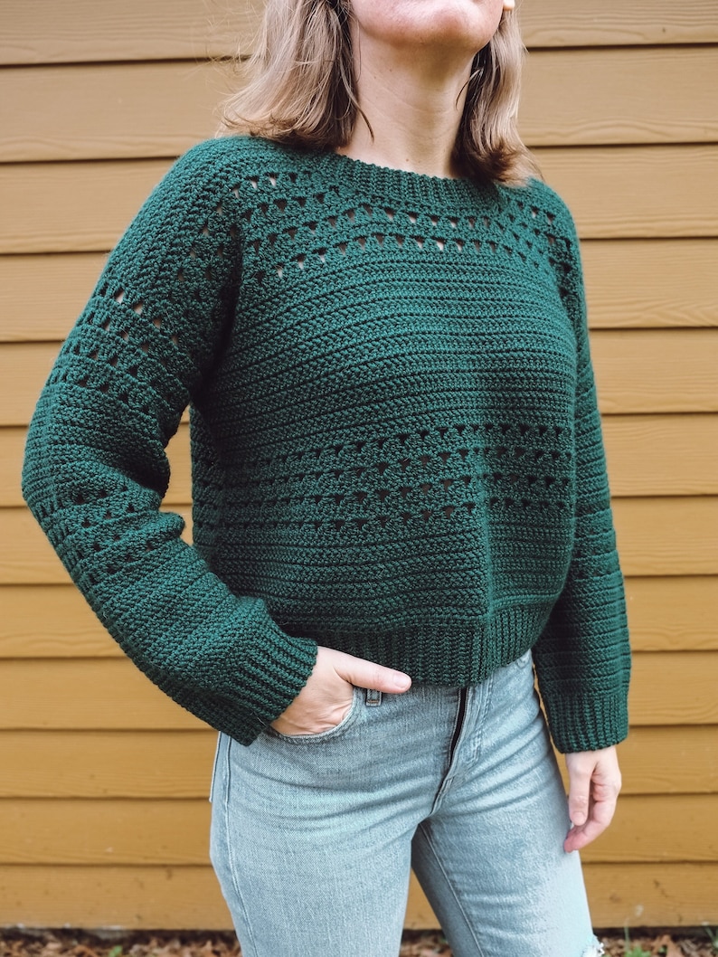 May include: A dark green crocheted sweater with a loose fit and a round neckline. The sweater has a lacy pattern throughout.
