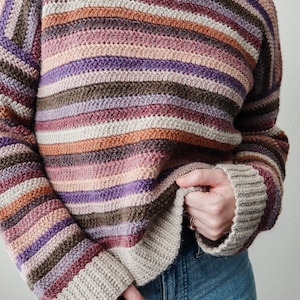Pu&ograve; includere: Un maglione a righe lavorato a uncinetto con una variet&agrave; di colori, tra cui viola, marrone, beige e rosa. Il maglione ha una vestibilit&agrave; rilassata e maniche lunghe.