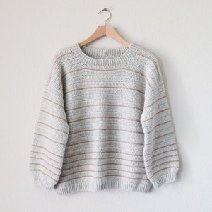 May include: A striped jumper in shades of grey and beige. The jumper has a round neckline and long sleeves.