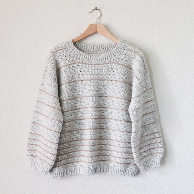 May include: A striped jumper in shades of grey and beige. The jumper has a round neckline and long sleeves.