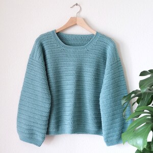 May include: A light blue crocheted sweater with long sleeves and a round neckline. The sweater is hanging on a wooden hanger against a white wall.