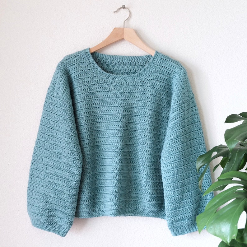 May include: A light blue crocheted sweater with long sleeves and a round neckline. The sweater is hanging on a wooden hanger against a white wall.