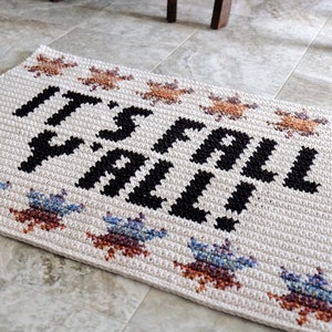 Puede incluir: Una alfombra de bienvenida blanca de crochet con el texto "It's Fall Y'all!" en negro. La alfombra tiene un borde de estrellas de crochet de colores.