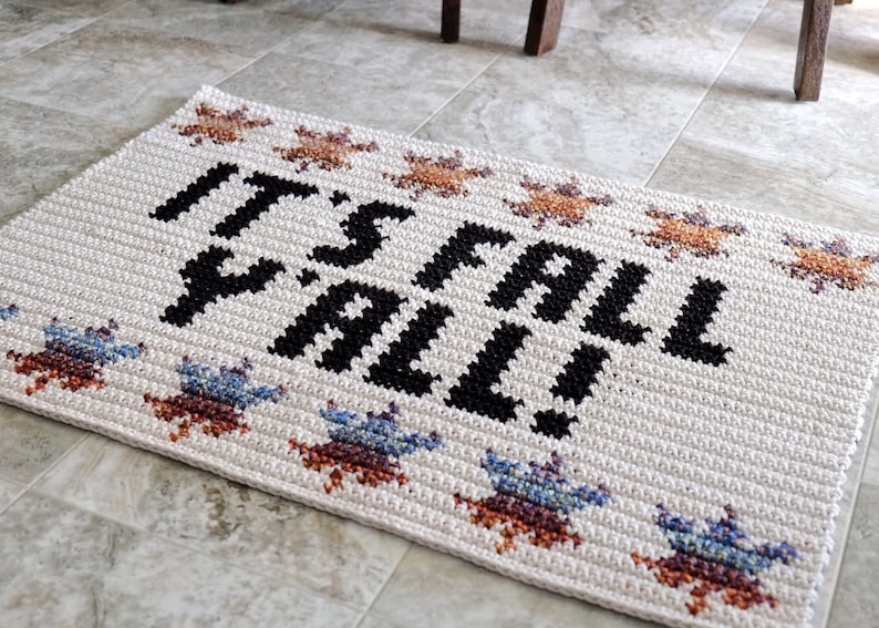 Puede incluir: Una alfombra de bienvenida blanca de crochet con el texto "It's Fall Y'all!" en negro. La alfombra tiene un borde de estrellas de crochet de colores.