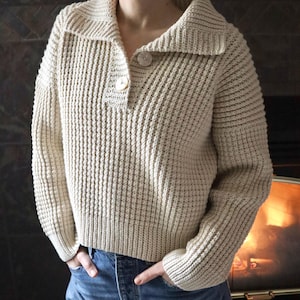 May include: A cream-colored knitted sweater with a collared neckline and two buttons. The sweater has a waffle knit texture and long sleeves.