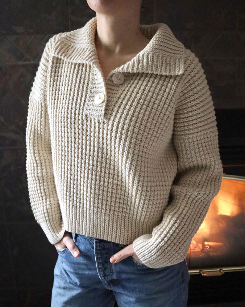 May include: A cream-colored knitted sweater with a collared neckline and two buttons. The sweater has a waffle knit texture and long sleeves.