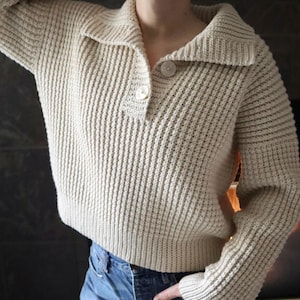 May include: A cream-colored knitted sweater with a collared neckline and a single button closure. The sweater has a ribbed texture and long sleeves.