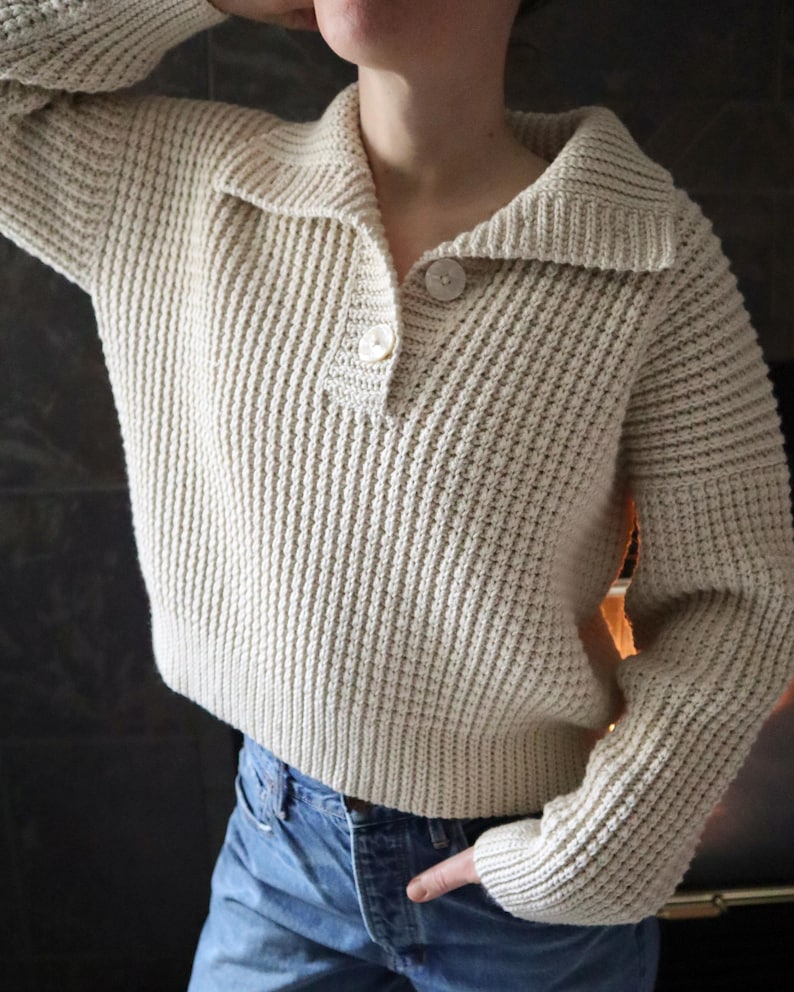 May include: A cream-colored knitted sweater with a collared neckline and a single button closure. The sweater has a ribbed texture and long sleeves.