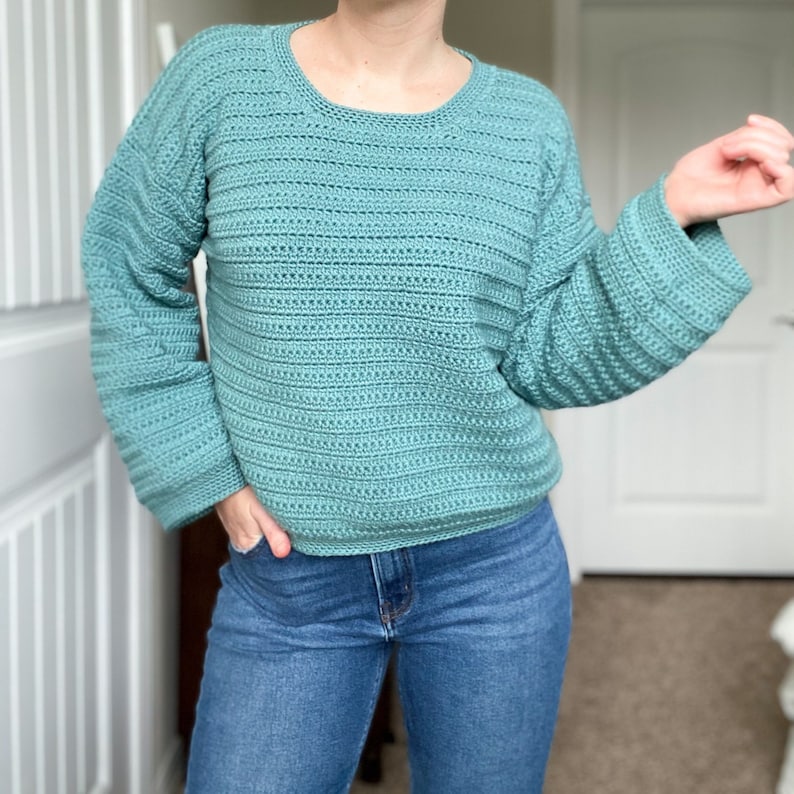 May include: A teal blue crocheted sweater with a round neckline and long sleeves. The sweater is made with a textured stitch and has a relaxed fit.