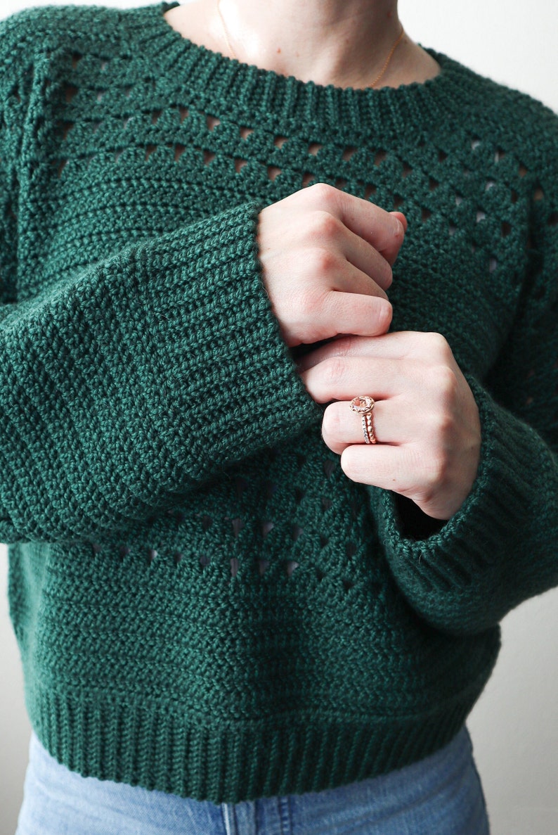 May include: A dark green crocheted sweater with a lacy pattern around the neckline and sleeves. The sweater is cropped and has a relaxed fit.
