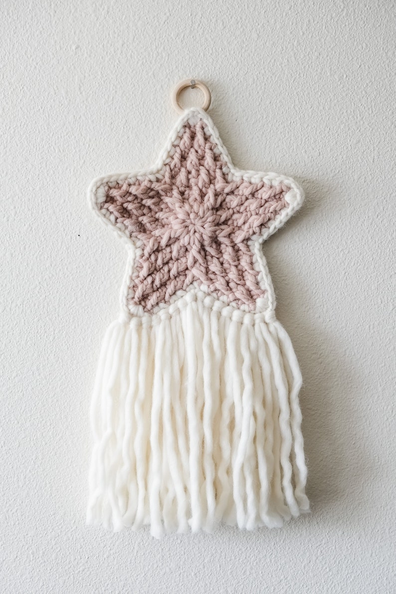 May include: A pink and white crocheted star with a white fringe hanging from a wooden ring.