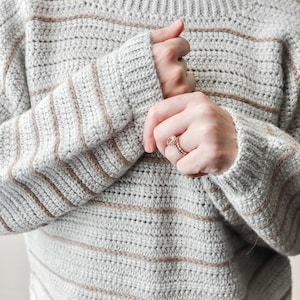 May include: A striped crocheted jumper in shades of grey and beige. The jumper has a relaxed fit and a round neckline.