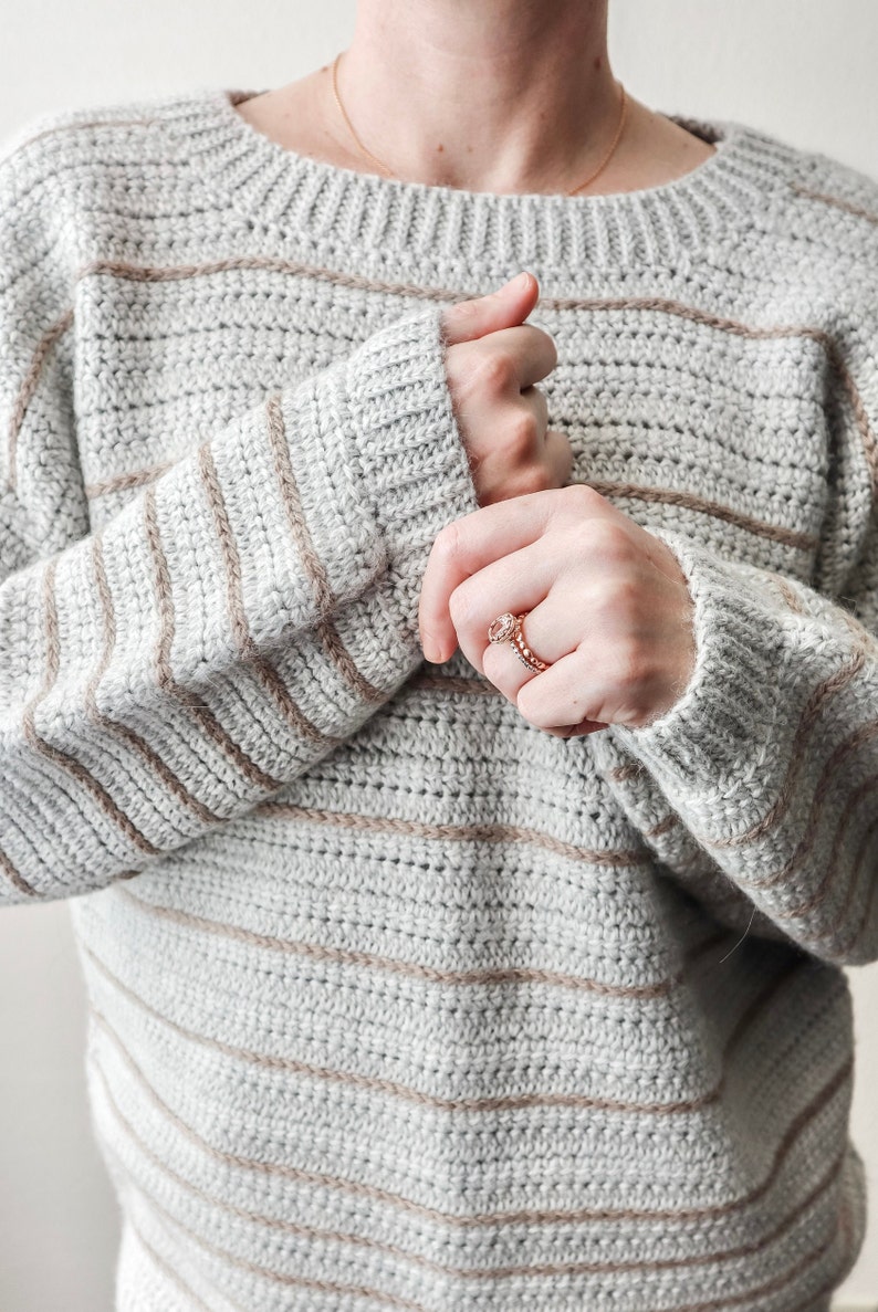May include: A striped crocheted jumper in shades of grey and beige. The jumper has a relaxed fit and a round neckline.