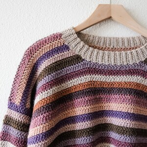 Pu&ograve; includere: Un maglione a righe lavorato a uncinetto in tonalit&agrave; di marrone, viola e rosa, appeso a un appendiabiti di legno su uno sfondo bianco.