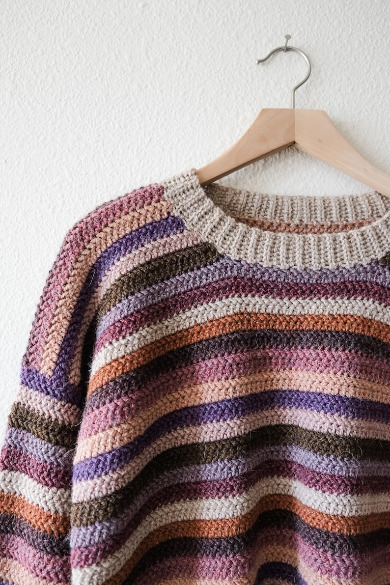 Pu&ograve; includere: Un maglione a righe lavorato a uncinetto in tonalit&agrave; di marrone, viola e rosa, appeso a un appendiabiti di legno su uno sfondo bianco.