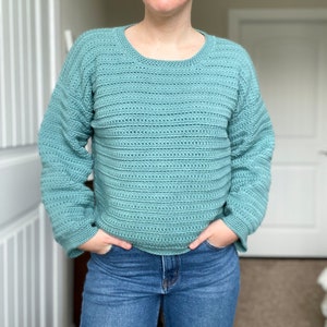 May include: A light blue crocheted sweater with a round neckline and long sleeves. The sweater is made with a textured stitch that creates a ribbed effect.
