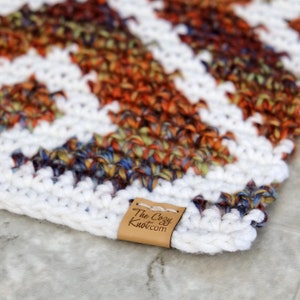 Może przedstawiać: Biały dywan wykonany na szydełku z wielokolorowym wzorem chevron. Dywan ma brązową sk&oacute;rzaną metkę z tekstem "The Cozy Knot.com".