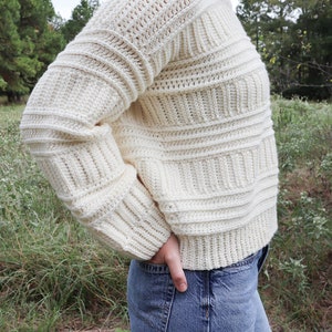 K&ouml;nnte beinhalten: Ein wei&szlig;er H&auml;kelpullover mit Rippenmuster. Der Pullover wird von einer Person mit blauer Jeans getragen.