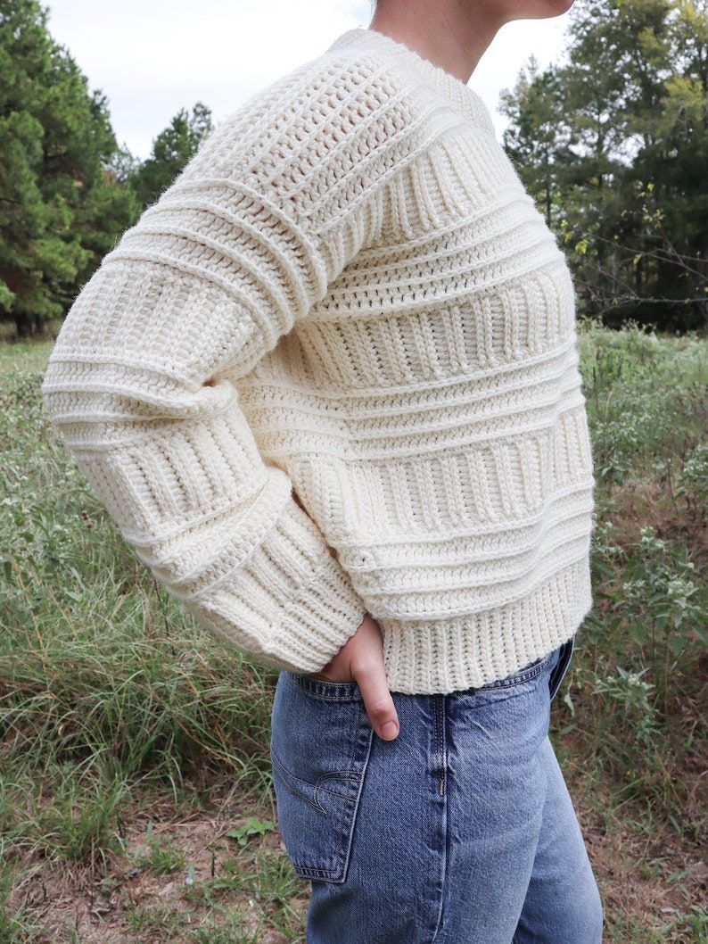 K&ouml;nnte beinhalten: Ein wei&szlig;er H&auml;kelpullover mit Rippenmuster. Der Pullover wird von einer Person mit blauer Jeans getragen.