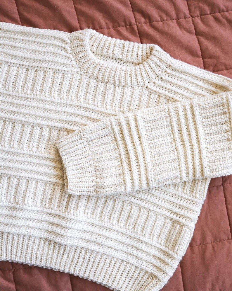 K&ouml;nnte beinhalten: Ein cremefarbener Strickpullover mit Rippenstruktur. Der Pullover hat einen Rundhalsausschnitt und lange &Auml;rmel.