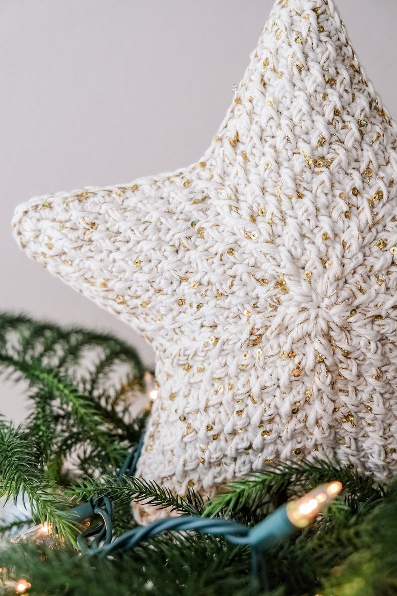 May include: A white crocheted star with gold sequins, used as a Christmas tree topper. The star is sitting on top of a green Christmas tree with lights.