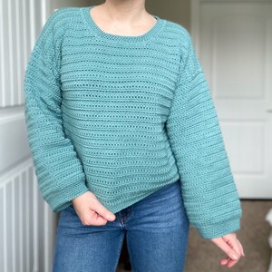 May include: A teal blue crocheted sweater with a loose fit and long sleeves. The sweater has a simple, open stitch pattern.