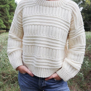 K&ouml;nnte beinhalten: Ein wei&szlig;er H&auml;kelpullover mit Rippenmuster und langen &Auml;rmeln. Der Pullover wird von einer Person mit einer blauen Jeans getragen.