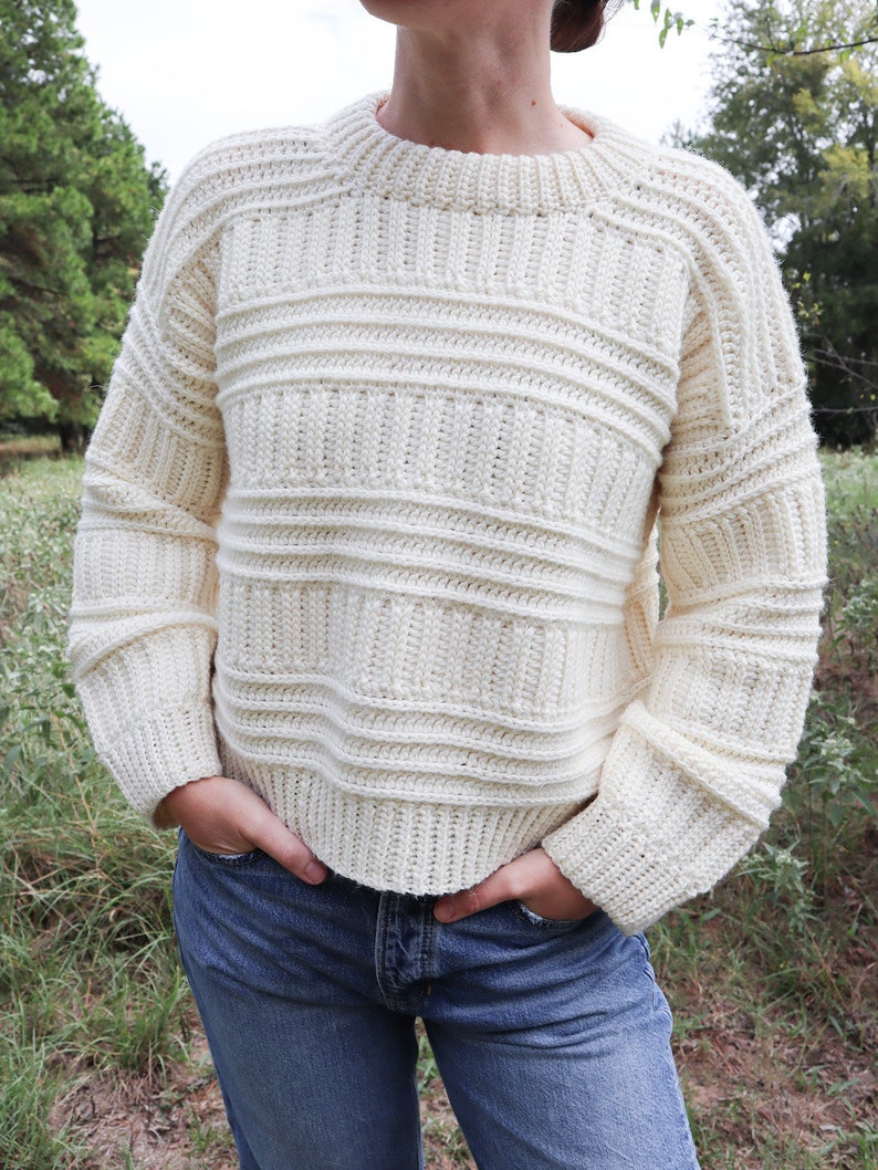 K&ouml;nnte beinhalten: Ein wei&szlig;er H&auml;kelpullover mit Rippenmuster und langen &Auml;rmeln. Der Pullover wird von einer Person mit einer blauen Jeans getragen.