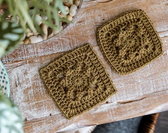 May include: Two crocheted square coasters in a muted olive green color. The coasters are made with a simple, repeating pattern and have a slightly textured surface.