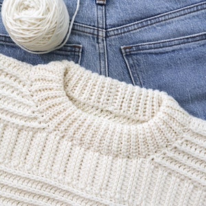K&ouml;nnte beinhalten: Ein wei&szlig;er H&auml;kelpullover mit einem strukturierten Muster, der auf einer blauen Jeanshose liegt. Eine wei&szlig;e Wollkn&auml;uel liegt auf der Jeans.