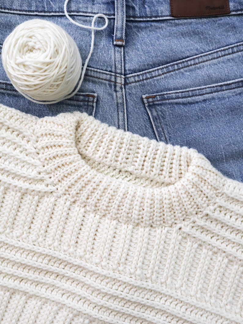 K&ouml;nnte beinhalten: Ein wei&szlig;er H&auml;kelpullover mit einem strukturierten Muster, der auf einer blauen Jeanshose liegt. Eine wei&szlig;e Wollkn&auml;uel liegt auf der Jeans.