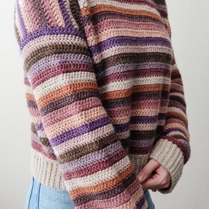 Pu&ograve; includere: Un maglione a righe lavorato a uncinetto in tonalit&agrave; di marrone, viola, rosa e beige. Il maglione ha un collo rotondo e maniche lunghe.