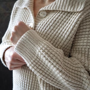 May include: A cream-colored knitted cardigan with a button-up front and a wide collar. The cardigan has a textured knit pattern and is made of a soft, cozy fabric.