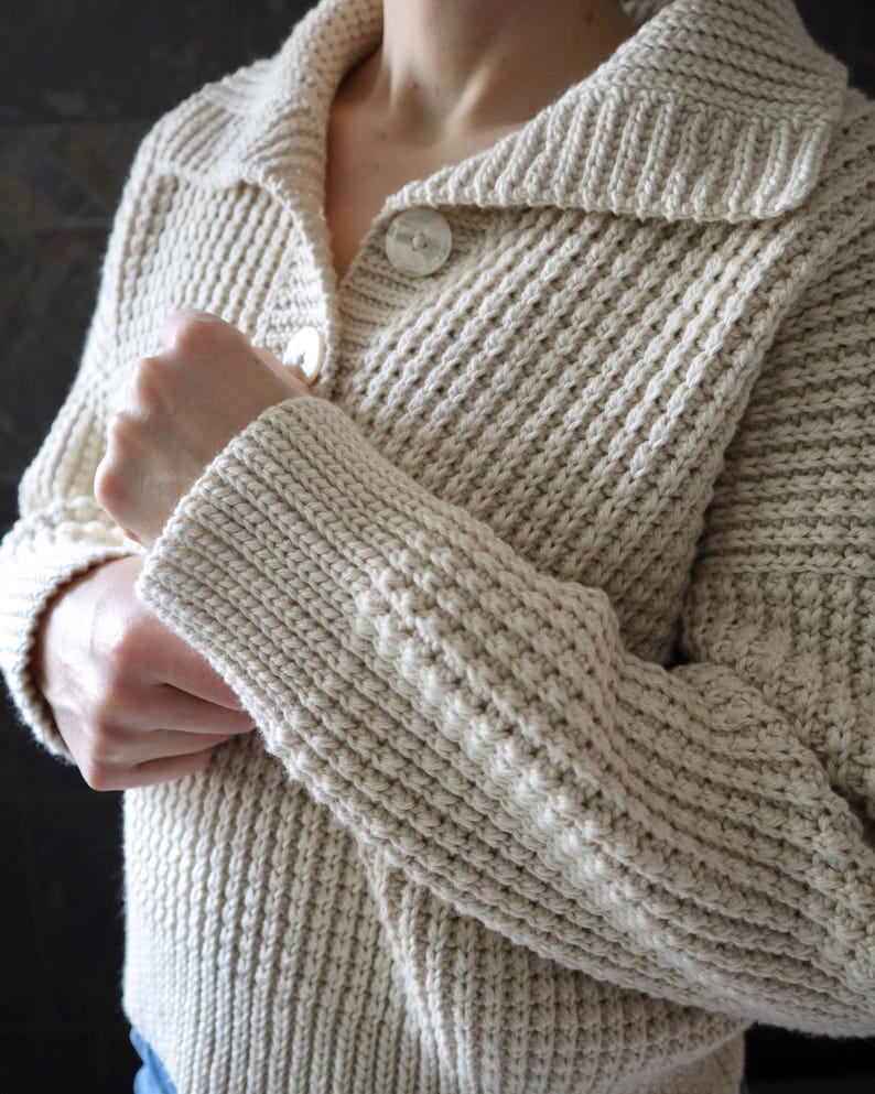 May include: A cream-colored knitted cardigan with a button-up front and a wide collar. The cardigan has a textured knit pattern and is made of a soft, cozy fabric.