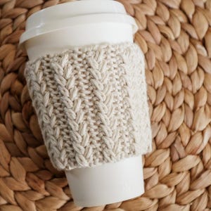 May include: A white takeaway coffee cup with a white lid and a cream-coloured knitted cosy. The cosy has a cable knit design. The cup and cosy are on a woven, light brown surface.