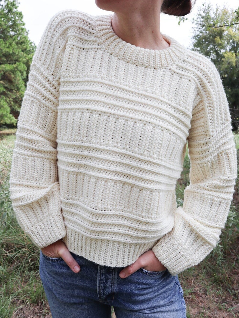 K&ouml;nnte beinhalten: Ein cremefarbener H&auml;kelpullover mit Rundhalsausschnitt und langen &Auml;rmeln. Der Pullover hat eine gerippte Struktur und ist aus einem dicken Garn gefertigt.