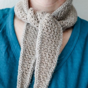 K&ouml;nnte beinhalten: Ein beigefarbener H&auml;kel-Schal, der um den Hals einer Person gebunden ist. Der Schal ist aus weichem, strukturiertem Garn gefertigt und hat ein schlichtes, elegantes Design.