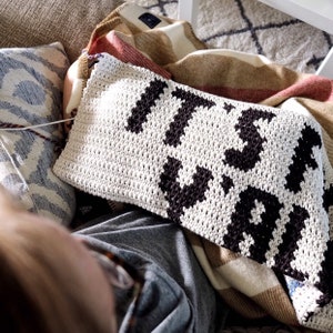 Puede incluir: Una manta de crochet blanca con letras negras que dicen "It's Wild". La manta est&aacute; colocada sobre un sof&aacute; con una persona sentada en &eacute;l.