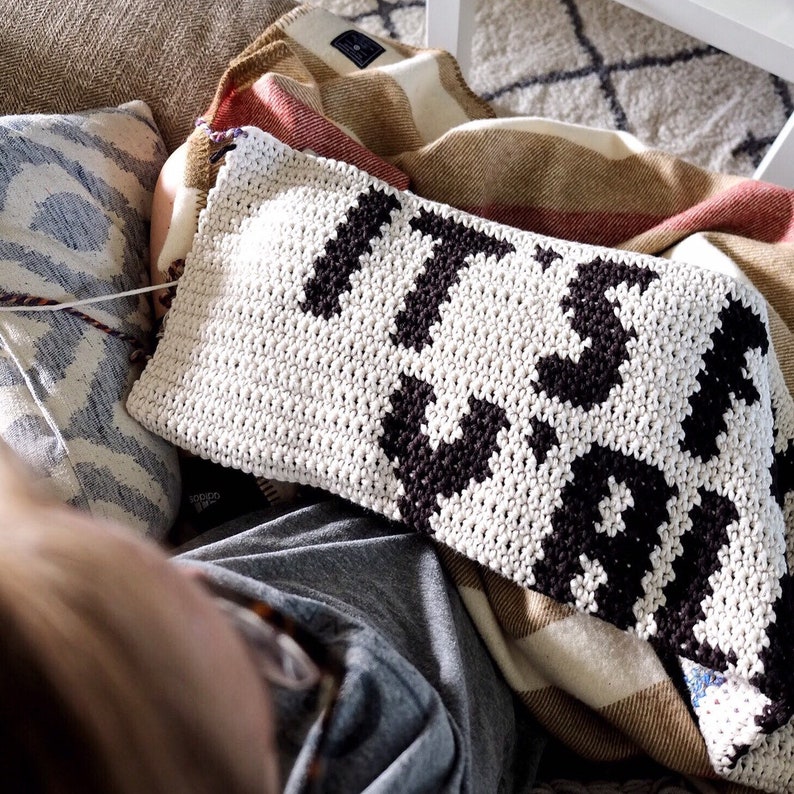 Puede incluir: Una manta de crochet blanca con letras negras que dicen "It's Wild". La manta est&aacute; colocada sobre un sof&aacute; con una persona sentada en &eacute;l.
