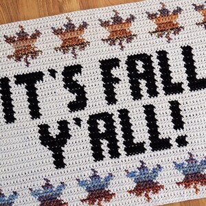 Puede incluir: Alfombra de crochet blanca con el texto "It's Fall Y'all!" en negro. La alfombra tiene un borde de hojas marrones y azules.