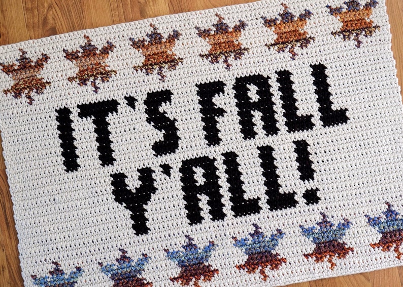 Puede incluir: Alfombra de crochet blanca con el texto "It's Fall Y'all!" en negro. La alfombra tiene un borde de hojas marrones y azules.