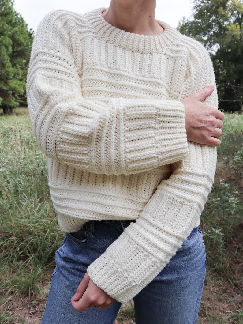 K&ouml;nnte beinhalten: Ein wei&szlig;er H&auml;kelpullover mit Rippenmuster und langen &Auml;rmeln. Der Pullover hat einen runden Ausschnitt und ist aus einem dicken Garn gefertigt.