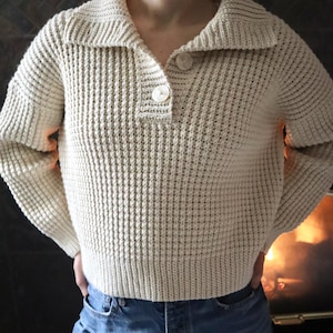 May include: A cream-colored waffle knit sweater with a collared neckline and a single button closure. The sweater has long sleeves and a relaxed fit.
