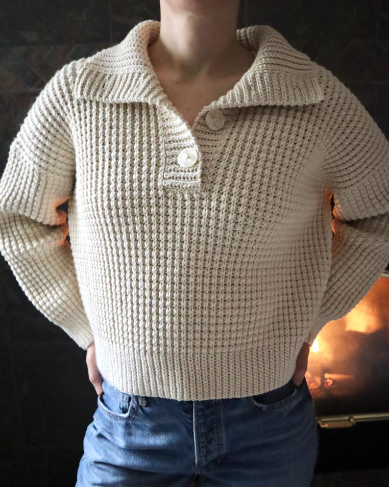 May include: A cream-colored waffle knit sweater with a collared neckline and a single button closure. The sweater has long sleeves and a relaxed fit.
