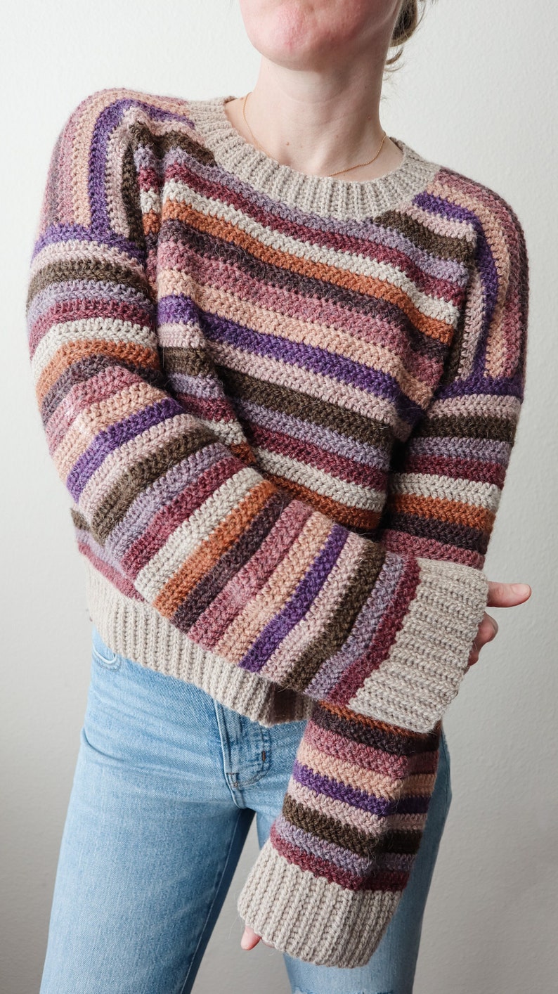 Pu&ograve; includere: Un maglione a righe lavorato a uncinetto con maniche lunghe in tonalit&agrave; di marrone, viola, rosa e beige.